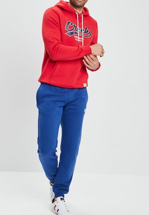 Sudadera roja con logo frontal, cordones blancos y pantalones de chándal azules. Presenta puños y cintura acanalados, con zapatillas blancas.