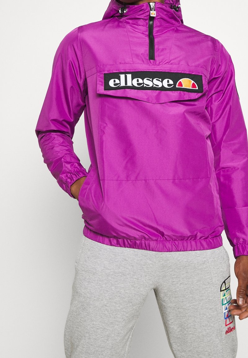 Fioletowa kurtka przeciwwiatrowa z przednią kieszenią i czarną naszywką z logo. Posiada regulowany kaptur i elastyczne mankiety dla lepszego dopasowania.