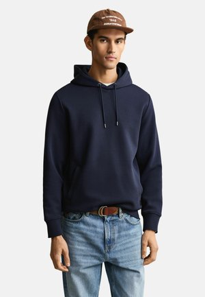 Jeune homme portant une casquette marron, un sweat à capuche bleu marine avec poche avant, un jean bleu clair et une ceinture marron, debout devant un fond uni.