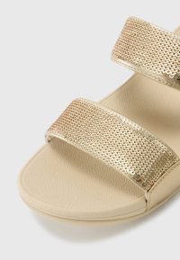 Gullfarget glidesandal med to brede teksturerte stropper, en glatt beige fotbed og design med rund tå.