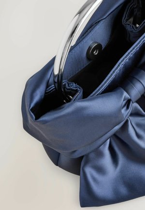 Sac à main en satin bleu marine avec un détail de nœud torsadé et une poignée en argent. L'intérieur présente un revêtement doux et une fermeture à pression magnétique.