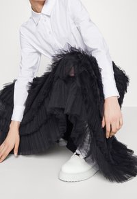 Gonna in tulle nera con plissee sovrapposte, abbinata a una camicia bianca con bottoni e scarpe platform bianche. La gonna presenta bordi volant.