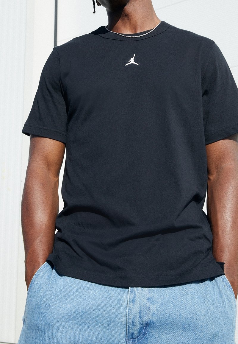 Schwarzes T-Shirt mit kurzen Ärmeln, das ein weißes Jumpman-Logo auf der Brust trägt, aus weichem Stoff gefertigt, kombiniert mit hellblauen Jeansshorts.