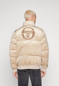 Giacca a vento beige realizzata in tessuto morbido, con colletto alto, polsini a costine e un grande logo ricamato sulla schiena.