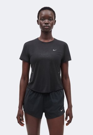 Kvinde står med fronten mod kameraet iført sort Nike sports t-shirt og shorts foran en ensfarvet lys baggrund.