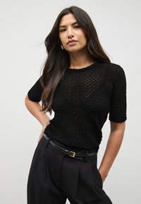 Top negro de manga corta con un patrón de punto texturizado, que presenta un escote redondo y se complementa con pantalones negros de talle alto y un cinturón.