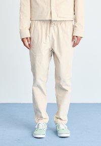 Beige bomulljacka med tryckknappar, kombinerad med byxor med elastisk midja. Modellen har på sig ljusgröna sneakers med vita sulor.