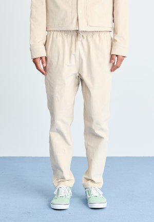 Veste en coton beige avec des boutons-pression, associée à un pantalon à taille élastique. Le modèle porte des sneakers vert clair avec des semelles blanches.
