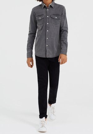 Child wearing a grey buttoned shirt, black pants, and white sneakers, standing against a plain light background.