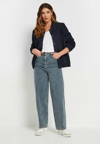 Marineblauwe gequiltte jas met knoopsluiting, witte V-hals T-shirt, hoge lichtblauwe spijkerbroek en witte sneakers met beige accenten.