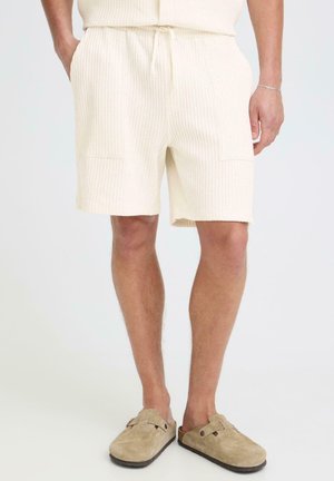 Mężczyzna w jasnobeżowych teksturowanych szortach z kieszeniami i dopasowanych beżowych butach wsuwanych, stojący na jednolitym tle.