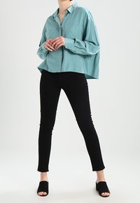 Camisa de botones en color turquesa con dos bolsillos frontales, corte holgado y mangas largas, combinada con pantalones negros ajustados y zuecos negros de punta abierta.