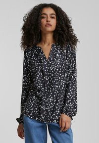 Svart langermet bluse med hvite blomster mønstre, V-hals, plisseringer foran og rynkede mansjetter. Brukt med lyseblå jeans.