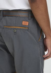 Gray shorts made of fabric featuring an orange accent stripe at the waistband, with a branded patch and a back pocket button detail.
