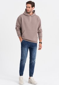 Haut à capuche beige avec une poche kangourou, associé à un jogging en denim bleu. Des baskets blanches et une montre noire complètent la tenue.
