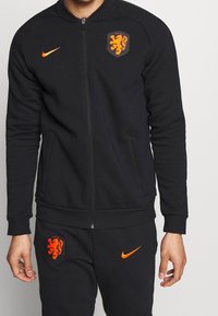 Zwarte Nike trainingspak met een jasje met ritssluiting en zakken. Beschikt over een oranje Nike swoosh en een geborduurd embleem op de borst en het broekbeen. Zachte stof.