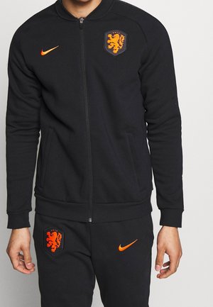 Tuta Nike nera con giacca con zip e tasche. Presenta lo swoosh arancione di Nike e uno stemma ricamato sul petto e sulla gamba del pantalone. Tessuto morbido.