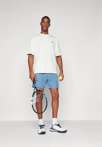 Světlounce šedé tričko, modré šortky, drží tenisovou raketu a míček. Sportovní boty s tmavými a bílými detaily. Jednoduchý design.