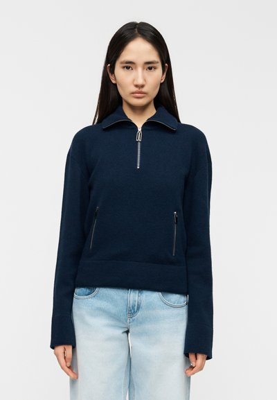 Pull-over en polaire bleu marine avec un col montant, une fermeture éclair sur la moitié de l'avant et deux poches zippées. Comprend des manches longues et une coupe décontractée.