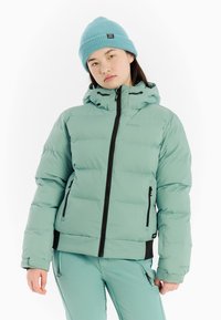Helles Teal-Puffer-Jacke mit schwarzem Reißverschluss, verstellbarem Kapuze und Seitentaschen. Kombiniert mit passenden Hosen und einer strukturierten Strickmütze.