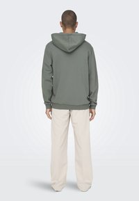 Sweat à capuche vert olive à coupe décontractée, avec poignets et ourlet côtelés, associé à un pantalon large couleur crème. Texture lisse, design décontracté.