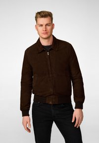 Brown suede bomber jacket with a collared neckline, zip front, and ribbed cuffs. Worn over a black top and paired with dark jeans.