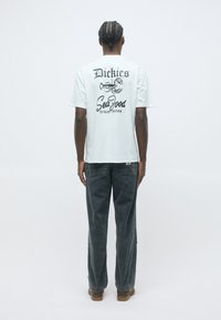 Man with braided hair wearing white graphic T-shirt with lobster and text, dark loose jeans, and brown shoes, standing against plain background.