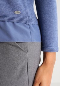 Blue knitted top with textured sleeves and a smooth lower panel, paired with gray pants featuring a side pocket detail.