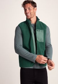 Smiling man adjusting sleeve wearing dark green fleece vest over gray long-sleeve shirt and black pants against plain background.