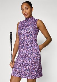 Bez piedurknēm violetas leoparda raksta kleita ar augstu apkakli, kas izgatavota no teksturēta auduma un ar sānu kabatām. Kreisajā rokā tur golfu nūju.