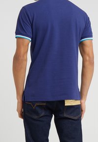 Polo shirt bleu marine au design ajusté et manches à rayures turquoise, associé à un jean en denim foncé avec un détail de poche arrière contrastant.