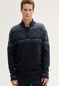 Granatowy sweter z dzianiny z wysokim kołnierzem, zamkiem błyskawicznym na pół, a górna część zdobiona jest wzorami geometrycznymi w jasnym niebieskim i szarym kolorze.