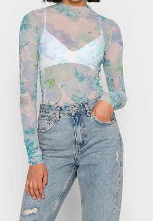 Femme portant un haut transparent à manches longues à motifs floraux dans des tons bleus et verts sur un bralette blanc, associé à un jean déchiré taille haute bleu clair.