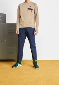 Osoba ubrana w beżowy długi rękaw Nike, ciemnoniebieskie spodnie i zielono-białe trampki Nike, stoi na musztardowym dywanie.