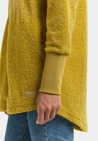Pull en polaire jaune avec une surface texturée et duveteuse, manches longues et une étiquette de marque à l'ourlet. Porté par-dessus une chemise à manches longues.