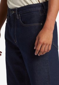 Mörkblå denimjeans med rak skärning, med klassisk femficksdesign, knappstängning och kontrasterande orange sömdetaljer.