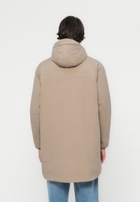 Beige gewatteerde jas met een capuchon, met lange mouwen en een verlengde achterkant. Gladde textuur, eenvoudig ontwerp en geen zichtbare hardware.