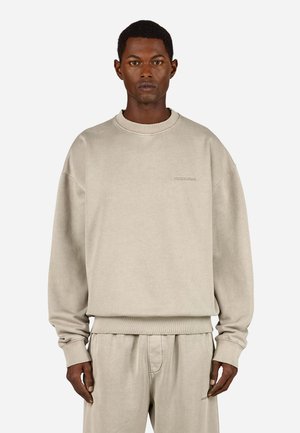 Ανοιχτό μπεζ φούτερ με στρογγυλή λαιμόκοψη, ριγωτές μανσέτες και ανάγλυφο λογότυπο στο στήθος, σε συνδυασμό με ταιριαστά παντελόνια.