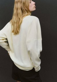 Kremowa bluza o luźnym kroju z opuszczonymi ramionami i miękką fakturą, zestawiona z czarnymi szortami na ciemnym tle.