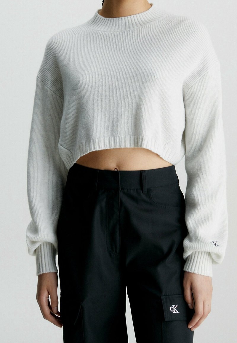Personne portant un pull court côtelé blanc et un pantalon noir taille haute avec les logos « cK » sur la manche et la poche.