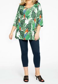 Tunique florale avec imprimé de feuilles de palmier vertes et de perroquets colorés. Encolure ronde, manches trois-quarts, associée à un jean en denim foncé et des mules noires.