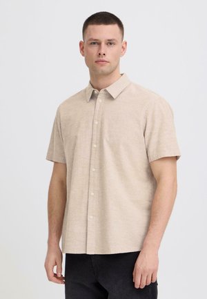 Młody mężczyzna stojący, ubrany w beżową, krótkorękawową koszulę na guziki oraz czarne spodnie, na tle jednolitego, jasnego tła.