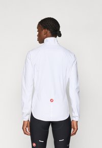 Weiße, leichte Radjacke mit hohem Kragen, elastischen Bündchen und einem reflektierenden Streifen auf dem Rücken. Mit einem roten Logo am Saum.