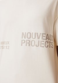 Helles beiges Baumwoll-T-Shirt mit Rundhalsausschnitt und kurzärmeligen Schnitt. Im Grauen aufgedruckter Text: "NOUVEAUX PROJECTS" und Akzente "75112".