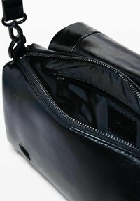 Sac en cuir noir à bandoulière avec fermeture éclair ouverte, montrant un intérieur lisse et une sangle réglable attachée avec un fermoir en métal noir.