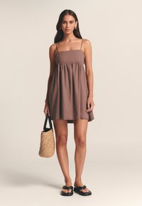 Abito in lino marrone con spalline sottili, vita arricciata e gonna svasata. Il modello tiene in mano una borsa intrecciata e indossa sandali neri.