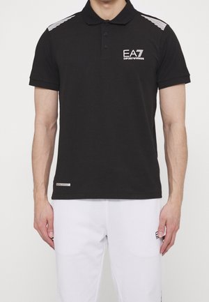 Mand iført en sort polo med hvide EA7 Emporio Armani-logoer på bryst og skuldre, kombineret med hvide bukser.