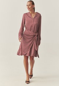 Rochie petrecută roșie cu model, mâneci lungi, guler și un detaliu cu nod lateral; țesătură netedă cu un model geometric circular.