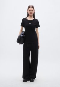 Črna majica s kratkimi rokavi in okroglim izrezom ter belim vezenim logotipom, kombinirana s širokimi črnimi hlačami in teksturirano črno torbico.