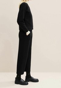 Personne debout de profil portant un haut à manches longues noir, un pantalon large noir, des chaussettes noires et des mocassins noirs sur un fond clair.